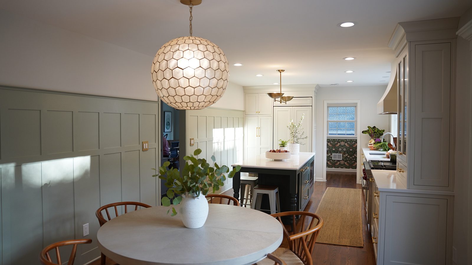 Dining table with wishbone chairs under a capiz-style globe pendant, with the green buffet and floral wallpaper behind