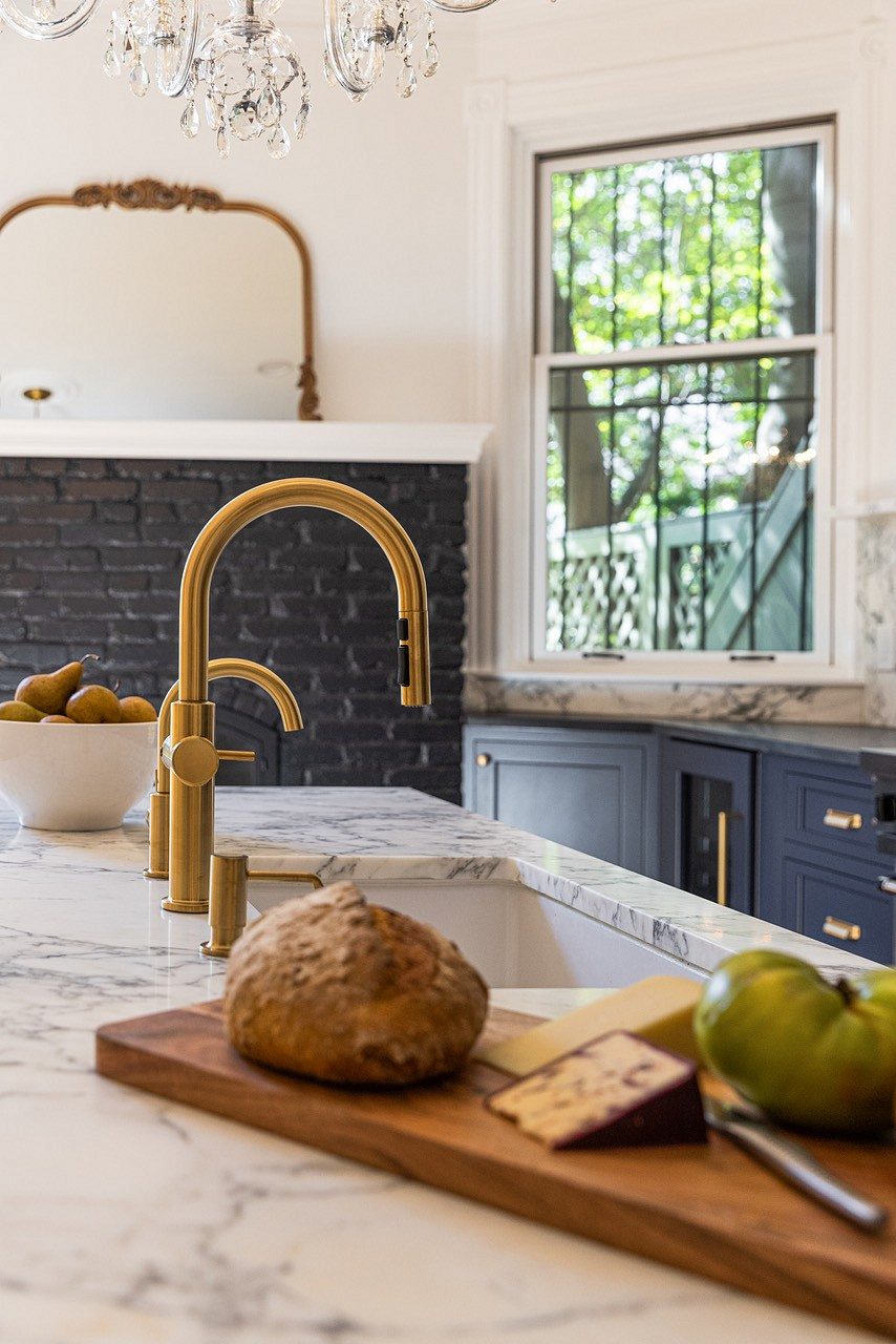 Brass faucets on the marble island top, with the black brick fireplace and bay window behind