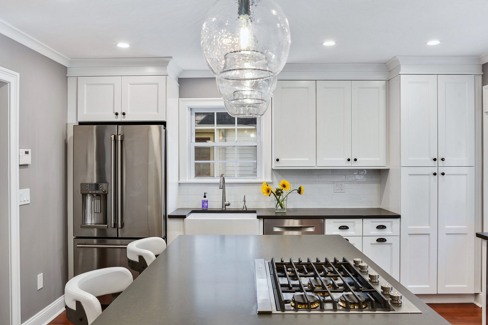 Gray island with a gas cooktop and white stools — Fabuwood white shaker kitchen with a gray island and glass pendants
