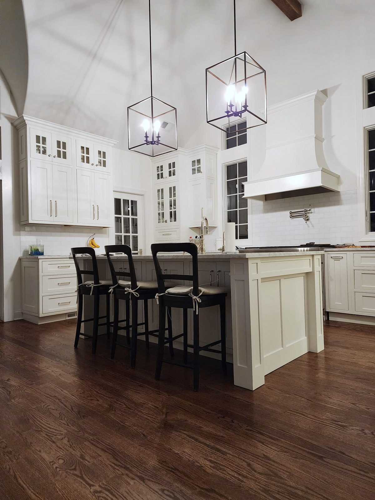 Marble island with a farmhouse sink and black stools — Shiloh Heritage beaded-inset kitchen under a vaulted ceiling with a curved hood