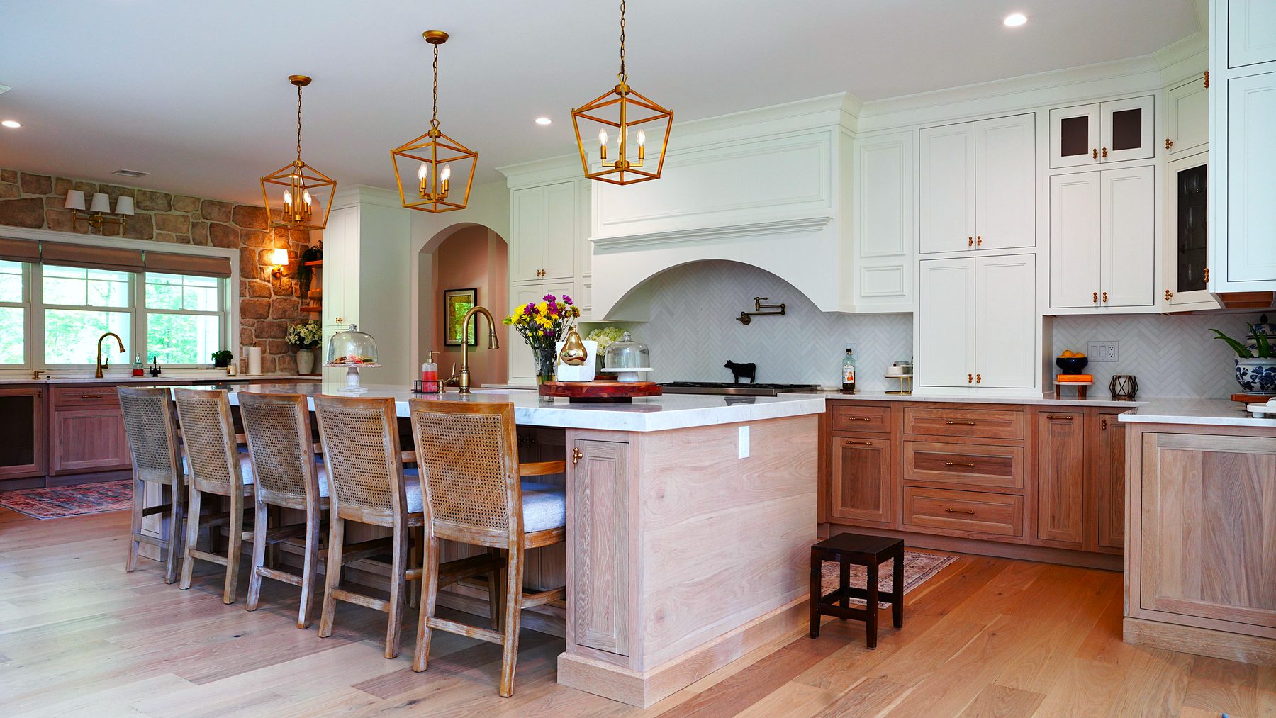 Two-tone kitchen with stained wood base cabinets, white uppers with glass panes, an arched plaster hood and brass lantern pendants over a long wood island