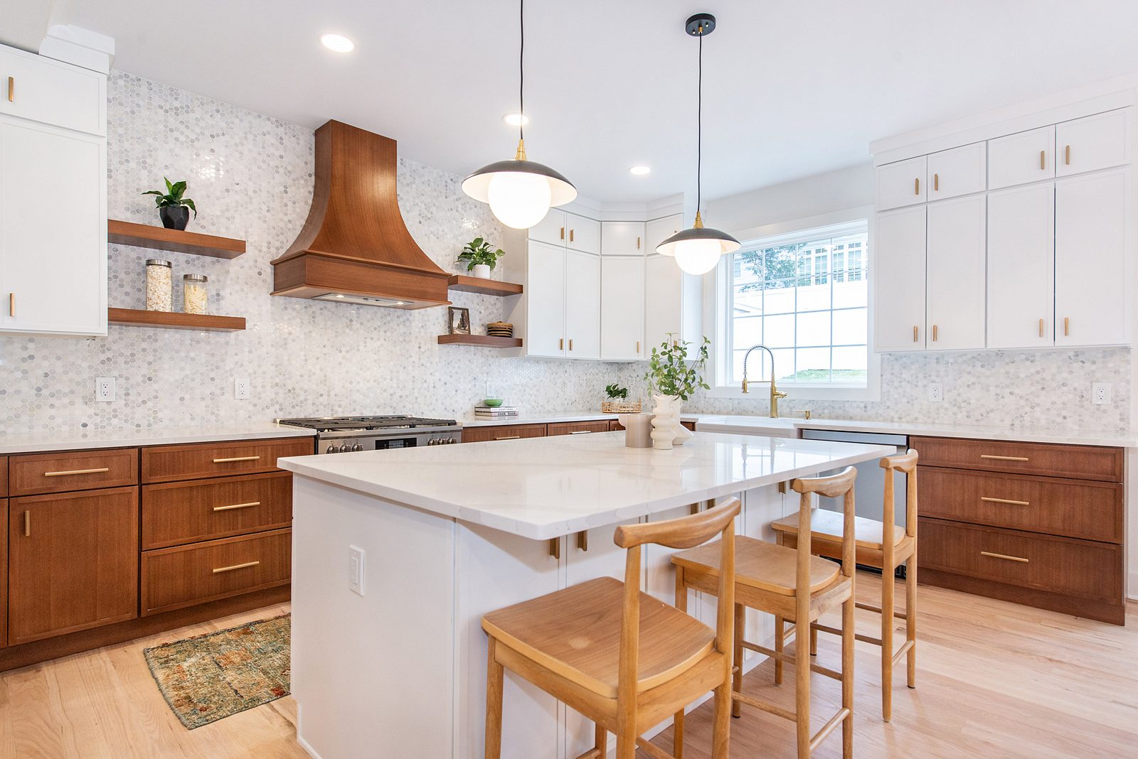 The range wall from the island: stained-wood hood between two floating shelves over a marble-mosaic backsplash