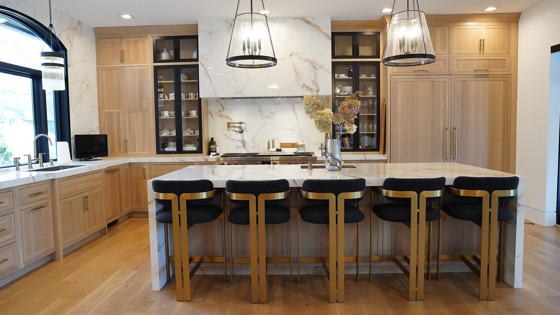 White oak kitchen with a Calacatta-look quartz island, six black-and-brass stools, drum pendants and an arched window